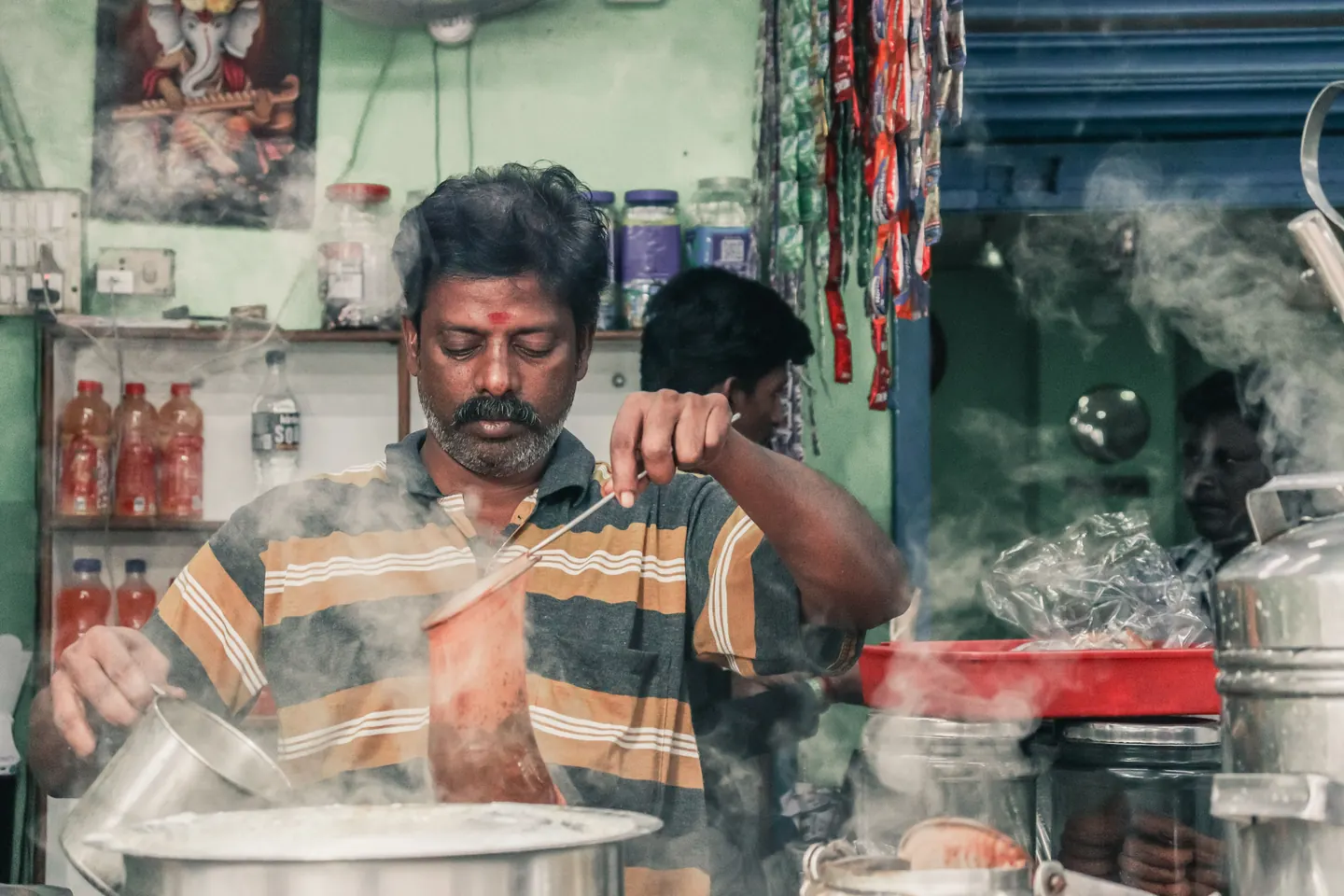 A tea seller pouring chai through a sieve at a market stall, steam rising from the pot.